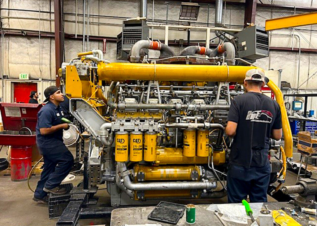 Two mechanics are working on a large yellow CAT industrial engine in a workshop. The engine is surrounded by various pipes, filters, and metal components, showcasing a complex system. One mechanic is standing on a step with tools in hand, inspecting the engine closely, while the other is working on the opposite side. The workshop environment includes equipment, toolboxes, and other industrial items. The setting is well-lit, with structural beams and electrical conduits visible in the background, contributing to the rugged, functional atmosphere.