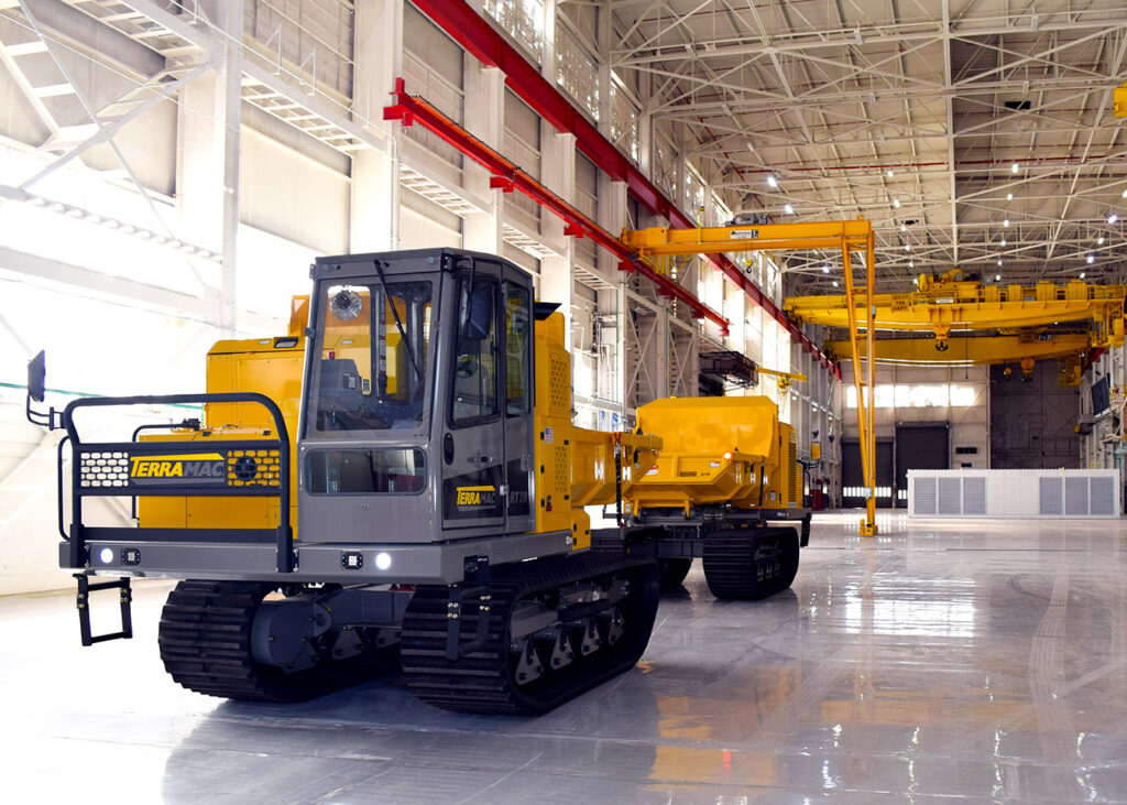 A yellow Terramac crawler is parked inside a spacious, well-lit manufacturing facility with a gleaming floor. The crawler has a rugged build with a cab and a heavy-duty track system, along with a trailer attached at the back. Overhead, large cranes are mounted on red and yellow rails, ready to assist with lifting and transporting heavy equipment. The expansive space and structured layout emphasize the scale and industrial capabilities of the facility, ideal for manufacturing or assembly processes.