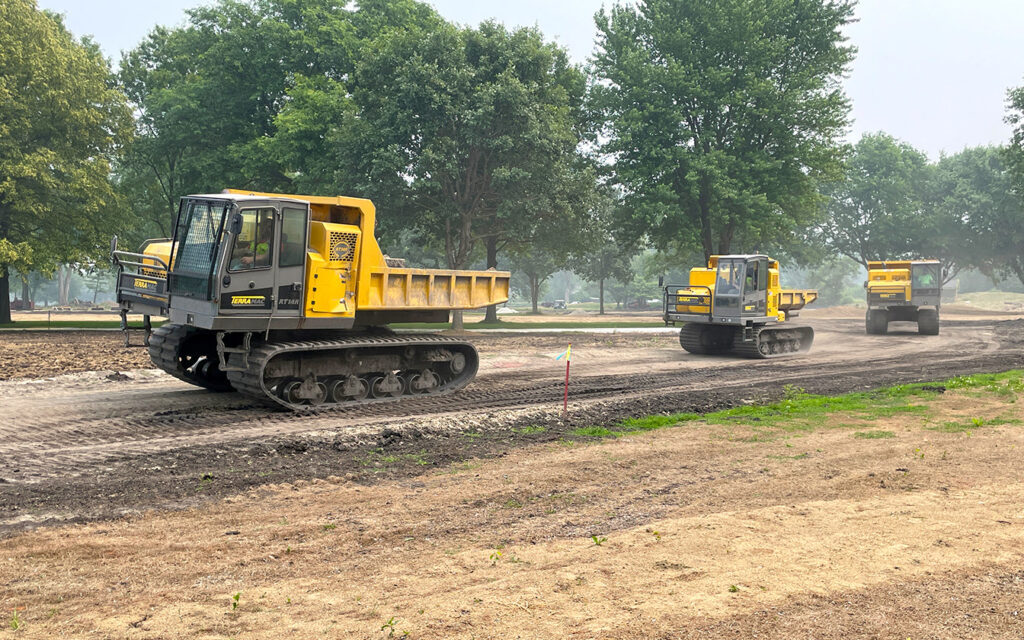 Three yellow Terramac crawler carriers drive on a construction site, surrounded by trees. The carriers, designed for heavy-duty off-road tasks, have large tracks for enhanced traction on uneven terrain. They are moving along a dirt path, preparing the site for further work amidst a landscape of grass and trees.