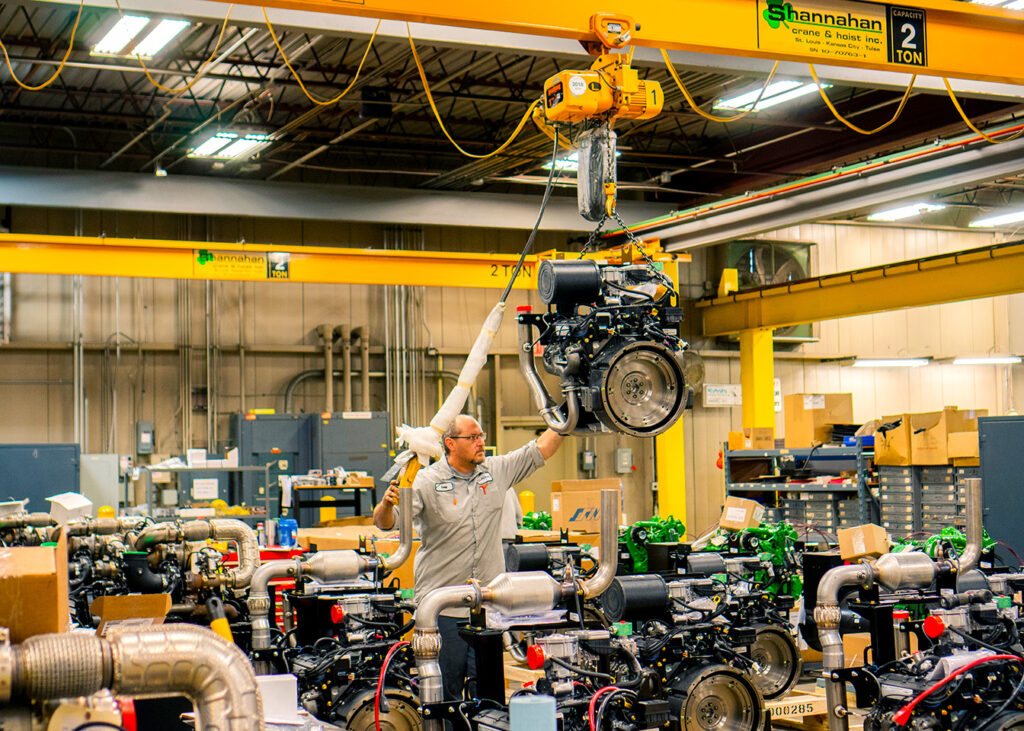 A worker in an industrial facility is operating a crane to lift a heavy engine part. The crane, marked with a 2-ton capacity sign, hangs from the ceiling and supports the engine as the worker guides it into position. The facility is filled with various engine components and machinery, with boxes and equipment spread throughout. Overhead lights illuminate the spacious workshop, and yellow support beams provide structural reinforcement.