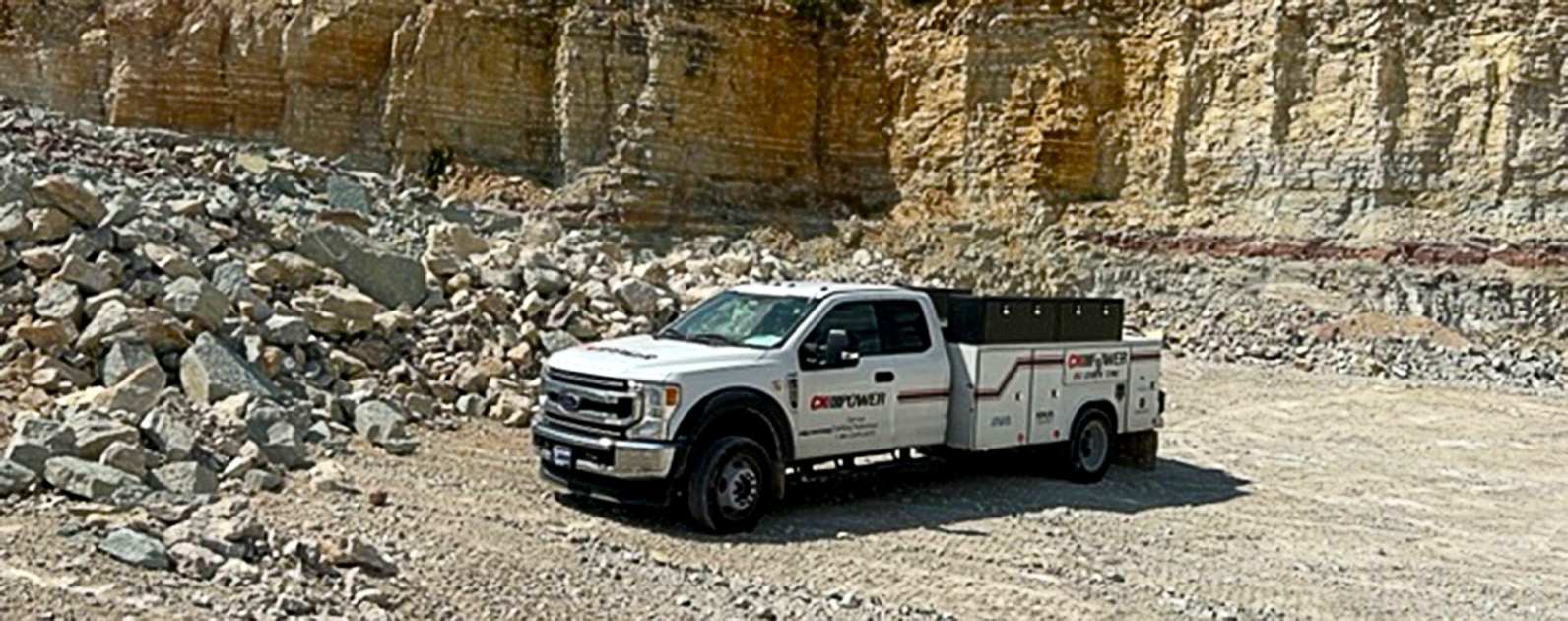 
A white CK Power field service truck is parked in a rugged quarry setting, surrounded by loose rocks and steep stone walls. The truck is equipped with tool compartments and appears ready for onsite maintenance work in challenging environments. The rocky landscape and exposed layers of sediment in the background highlight the remote and industrial nature of the location, emphasizing the truck’s capability to provide service in hard-to-reach areas.