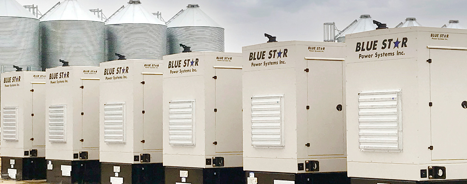 A row of generators stands aligned outdoors, with several large metallic silos in the background. The industrial setting under a cloudy sky creates a robust atmosphere, showcasing reliable power solutions for commercial or agricultural applications.