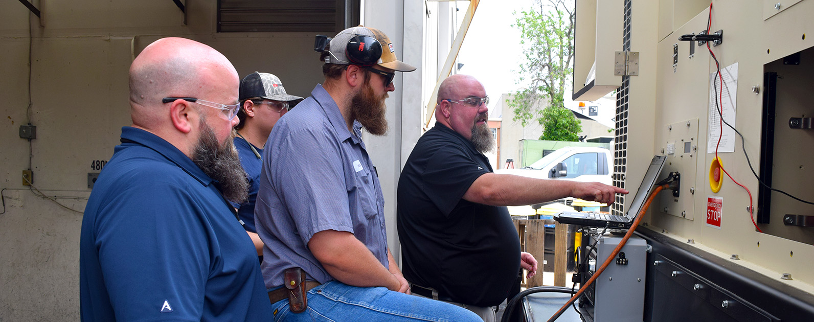 
Four men are gathered around industrial equipment, closely examining a control panel. One man, wearing a black shirt and safety glasses, is pointing at the panel, possibly explaining or demonstrating something to the group. The others, dressed in work shirts and protective gear, listen attentively. The equipment has emergency stop buttons and wiring, indicating a focus on safety and technical details. The outdoor setting suggests a hands-on training session or inspection, emphasizing teamwork and shared expertise in an industrial environment.