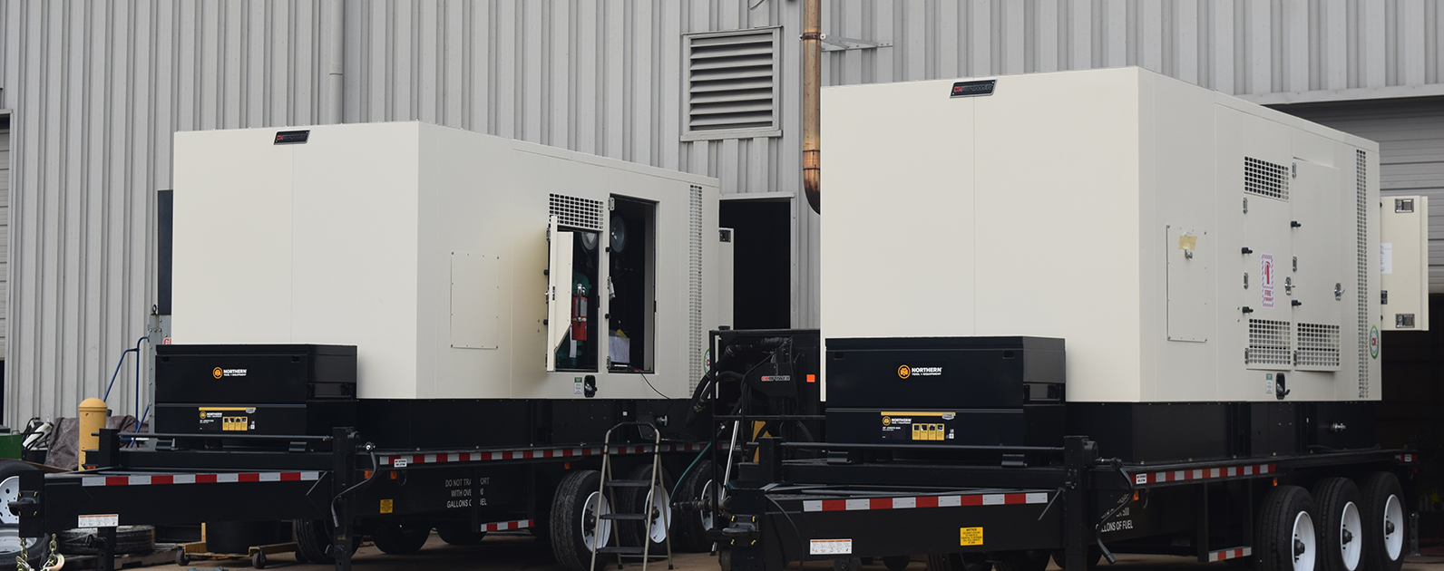 Two large industrial generators mounted on trailers outside a manufacturing facility. Each generator has a beige casing with ventilation grates and an open access panel displaying interior components. 