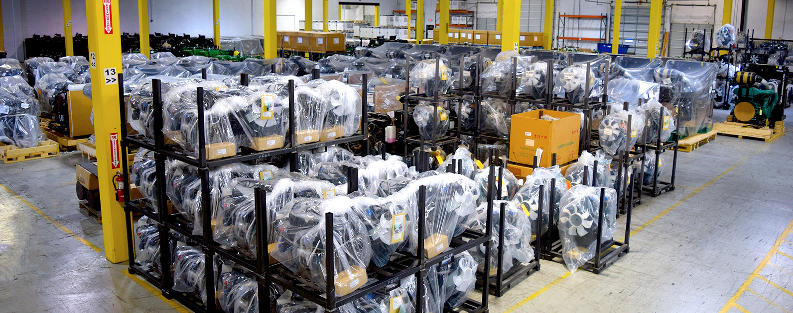 A warehouse filled with racks of engines neatly wrapped in plastic for protection. The engines are stacked on metal frames and organized in rows, ready for distribution. Yellow safety columns and marked pathways indicate a well-organized storage area within an industrial facility. Boxes and other equipment are visible among the rows, showcasing a busy inventory setting prepared to supply various off-highway equipment needs. The scene conveys efficiency and readiness in managing large-scale engine distribution.