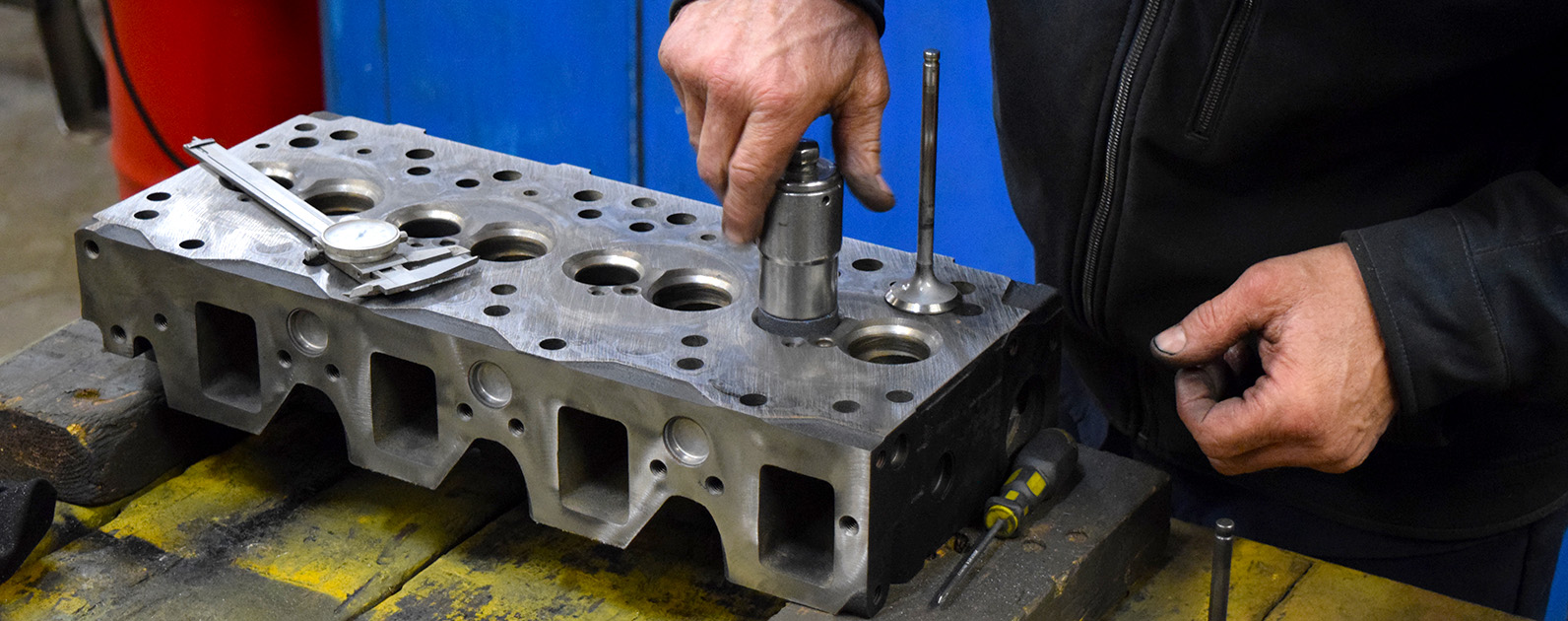 The image shows a close-up of a technician's hands working on a metal engine cylinder head in a repair shop. The technician is inserting a valve guide into one of the holes on the cylinder head, with a dial caliper placed nearby for precision measurement. Various other tools, including a screwdriver, are scattered around the workbench. The background includes a blue divider, suggesting a designated workspace within the shop. The scene emphasizes the careful, detailed work involved in rebuilding and repairing engine components, highlighting technical skill and accuracy.