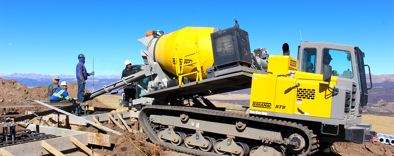 
A yellow Terramac RT9 rubber-tracked crawler carrier equipped with a large cement mixer is positioned on a rugged construction site with mountainous terrain in the background. Construction workers in hard hats and safety vests are nearby, guiding the concrete chute to pour cement into a foundation. The vehicle's durable tracks allow it to maneuver across the uneven ground, highlighting its versatility for off-road construction tasks in challenging environments.