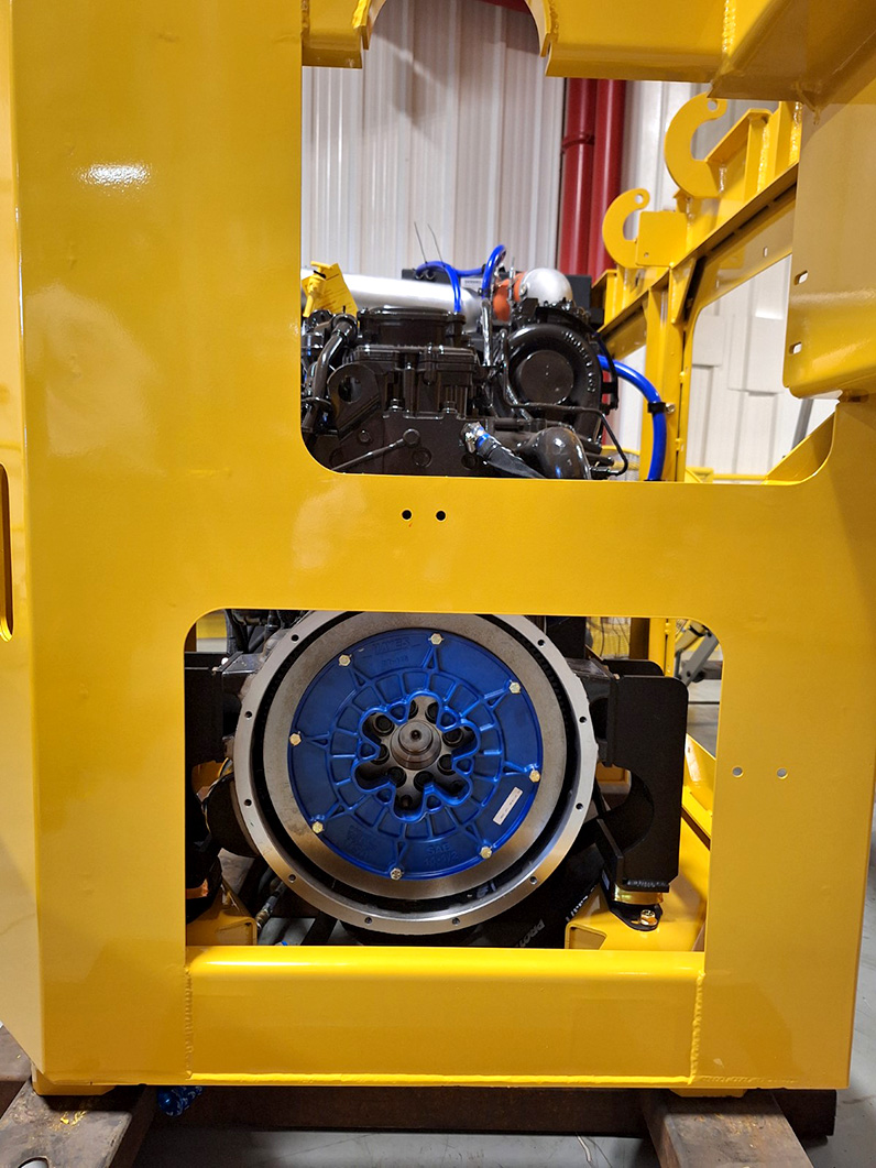 Close-up view of a Hayes coupling installed on a Terramac crawler carrier, housed within a bright yellow frame. The blue coupling and surrounding components contrast against the yellow, highlighting the precision-engineered connection between the engine and the machinery. The industrial setting in the background adds context to this robust piece of equipment.