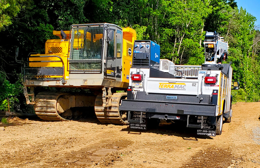 A yellow Terramac crawler carrier with protective metal grating on the cab is parked next to a white Terramac service truck on a dirt path. The crawler has heavy-duty tracks and appears rugged, designed for navigating rough terrain. The service truck is equipped with various tools, equipment, and a mounted crane, indicating it’s ready for field maintenance work. The scene is set against a backdrop of dense, green foliage, suggesting a remote or outdoor worksite. The dirt ground shows signs of recent activity, with tire tracks and compacted soil.