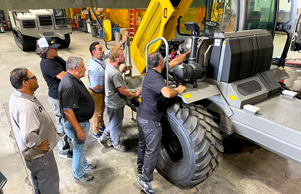 A group of six men is gathered around a large wheeled dumper in a workshop, closely examining its engine and components. Some are holding onto the equipment or leaning in to get a better view, indicating a training or troubleshooting session. They are dressed in a mix of casual and work attire, suggesting different roles or levels of expertise. The scene is set in a well-equipped workshop with various tools and machinery in the background, emphasizing hands-on learning and collaboration in an industrial setting.