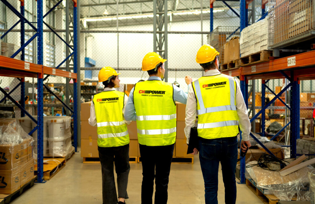 
Three people wearing yellow hard hats and CK Power-branded safety vests walk through a warehouse with shelving units and packed pallets.