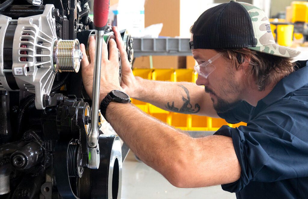 
A mechanic wearing safety glasses and a backward camo hat is intently working on an engine. He is using a wrench and appears focused on adjusting or securing a component. The mechanic's forearm tattoo and watch add a personal touch to his work attire. The background features organized bins, suggesting a well-equipped and orderly workshop. The scene captures the precision and concentration required in industrial equipment maintenance.