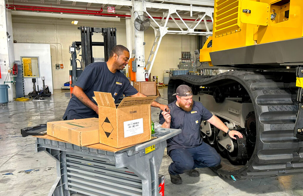 Two mechanics work together on a large rubber-tracked crawler carrier in a well-lit industrial facility. One stands next to a cart filled with boxes, looking at his colleague with a smile. The other mechanic is crouched down, inspecting the carrier's undercarriage and pointing at a specific part. They both wear matching navy work uniforms, appearing focused and engaged in their task.