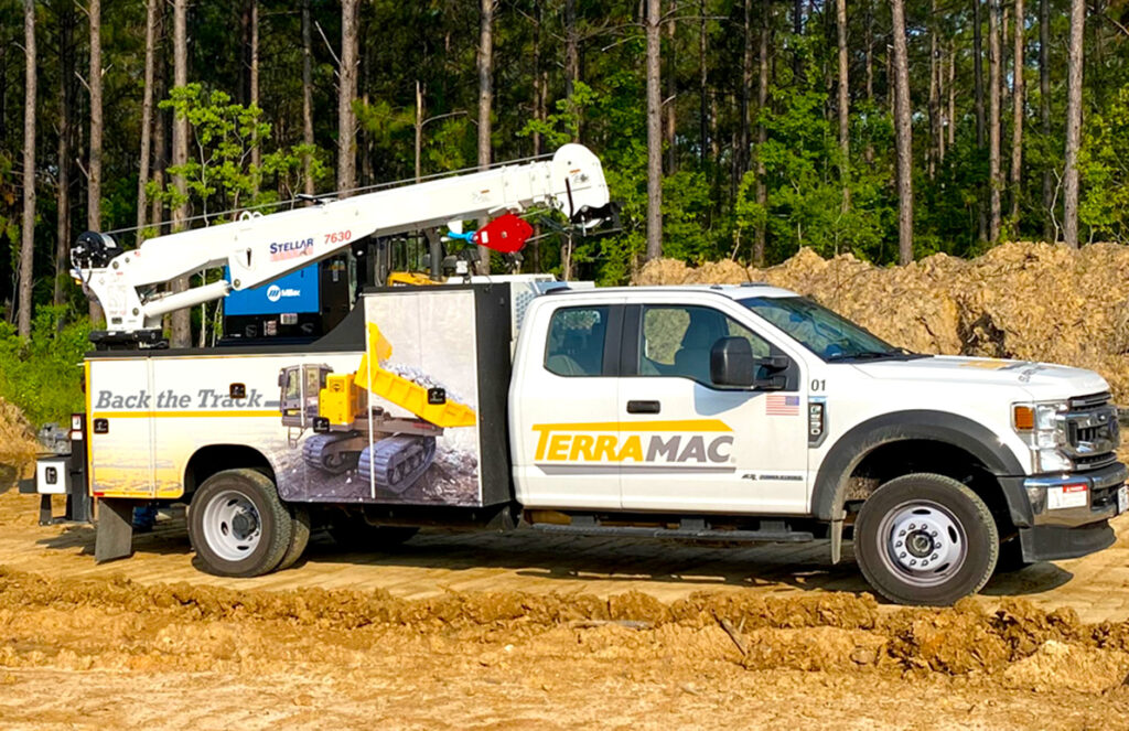 A white Terramac service truck with a mounted crane is parked on a dirt construction site, surrounded by a dense forest. The truck features branding on the doors and sides, showcasing an image of a yellow crawler carrier. The text "Back the Track" is visible on the side panel. The truck is equipped with tools and equipment for field maintenance, including a Stellar 7630 crane and a Miller welding unit, emphasizing its role in providing aftermarket support for heavy machinery.