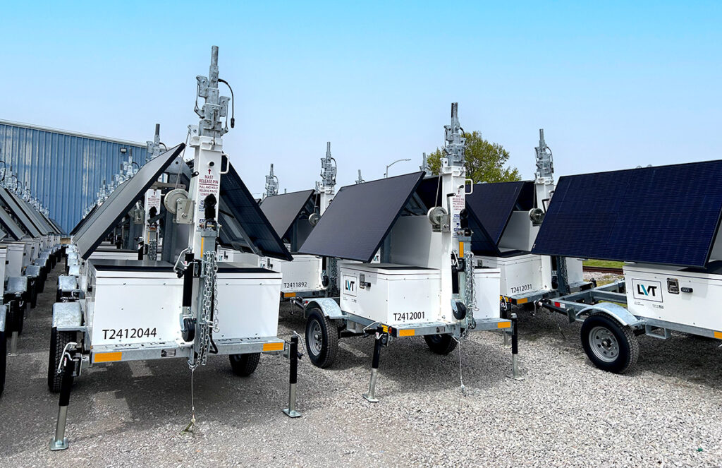 A row of portable solar-powered light towers, each equipped with large solar panels and extendable masts, is parked on a gravel lot. 