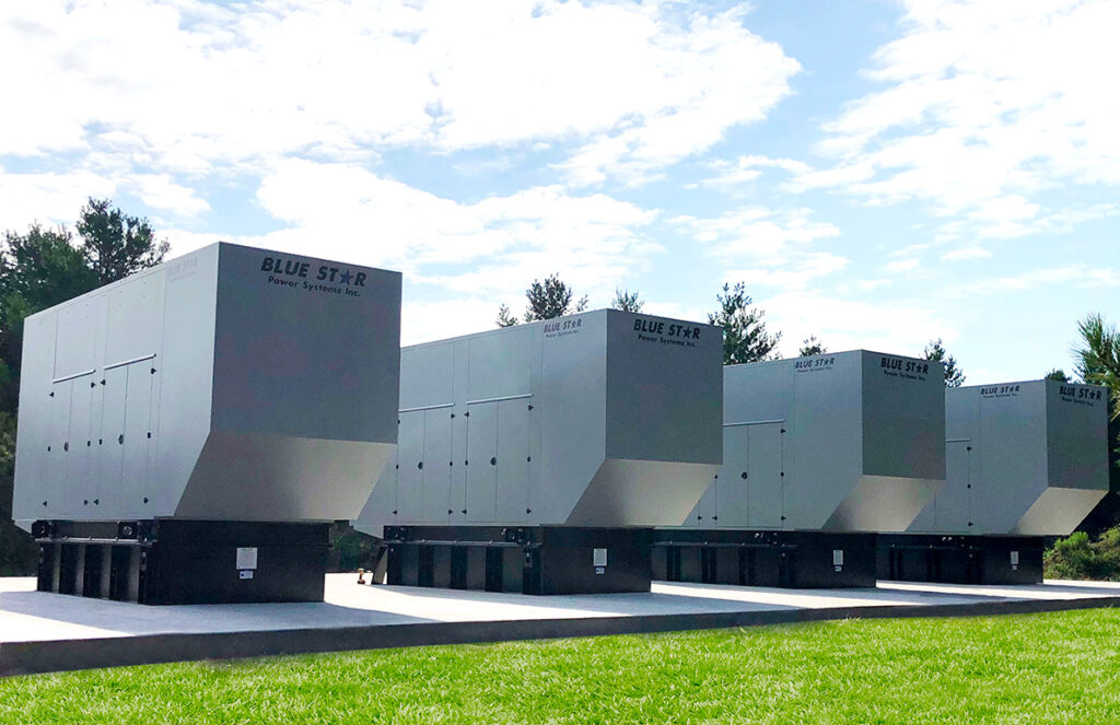 
A row of four large Blue Star Power Systems generators is lined up outdoors on concrete pads. The generators have sleek, rectangular enclosures and are elevated on sturdy black bases. The sky is partly cloudy, and the surrounding area is grassy with trees in the background, suggesting a serene, well-maintained installation site. The setup conveys a sense of reliability and readiness, with the generators positioned to provide backup power when needed.
