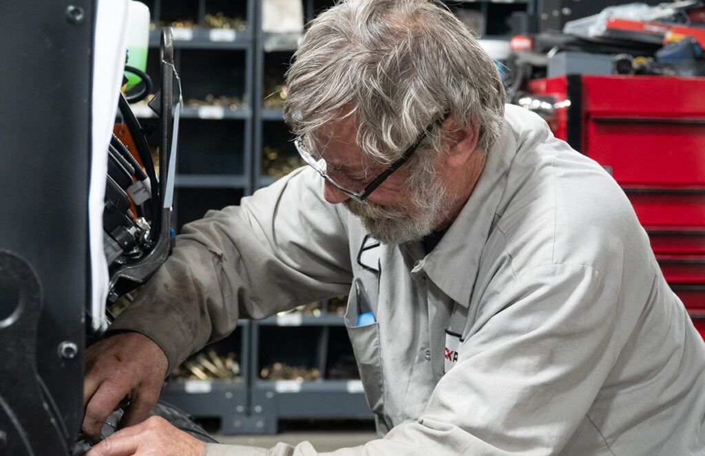 
An older mechanic with gray hair and a beard is working intently on a piece of equipment in a workshop. He is wearing safety glasses and a light gray work shirt. The mechanic's workspace is organized, with storage shelves filled with parts and a red toolbox visible in the background. The environment reflects a well-used and practical setup, suitable for detailed repair work.