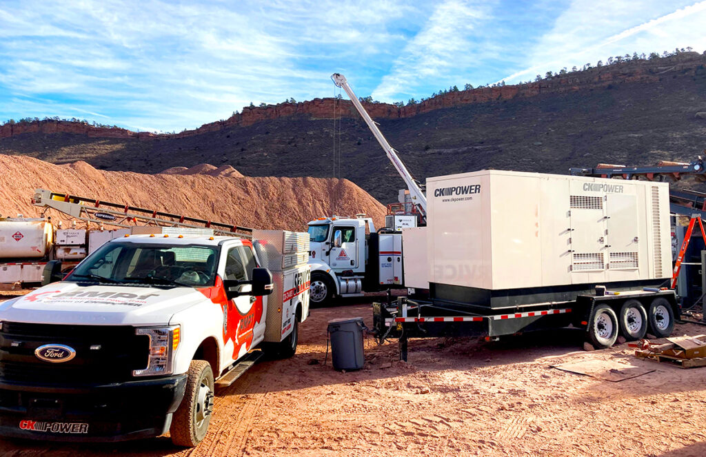 The image shows a service site in a rugged, outdoor setting with a large off-highway generator unit mounted on a trailer. A branded service truck, equipped with tools and supplies, is parked nearby, with another utility truck and fuel tanker visible in the background. The site is set against a backdrop of rocky hills under a clear blue sky. The scene illustrates on-site service and repair operations for heavy-duty power equipment, with the trucks and tools highlighting the mobile, field-based support necessary for off-highway OEM equipment.