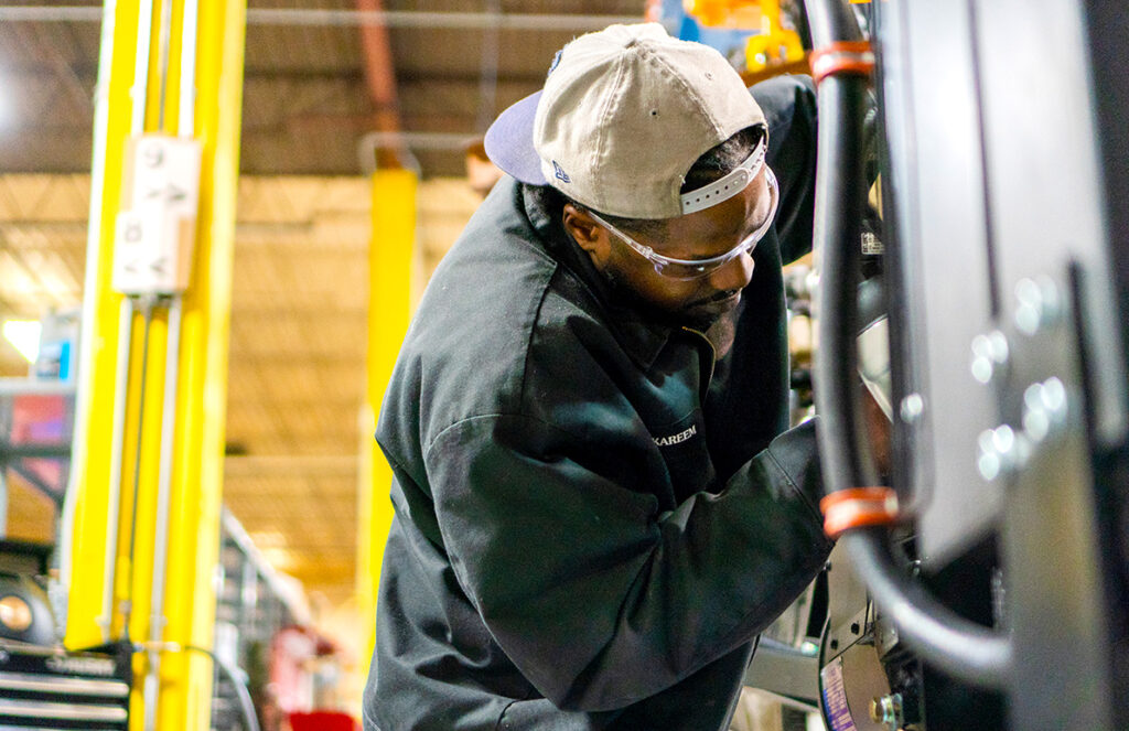 A worker wearing safety glasses and a baseball cap backward is focused on inspecting or repairing industrial equipment. He is dressed in a black jacket and leans in closely to examine the components, suggesting a hands-on approach. The brightly lit workshop has yellow support columns and various machinery, creating a busy and productive industrial environment. The scene highlights the attention to detail and technical skills involved in aftermarket industrial solutions.