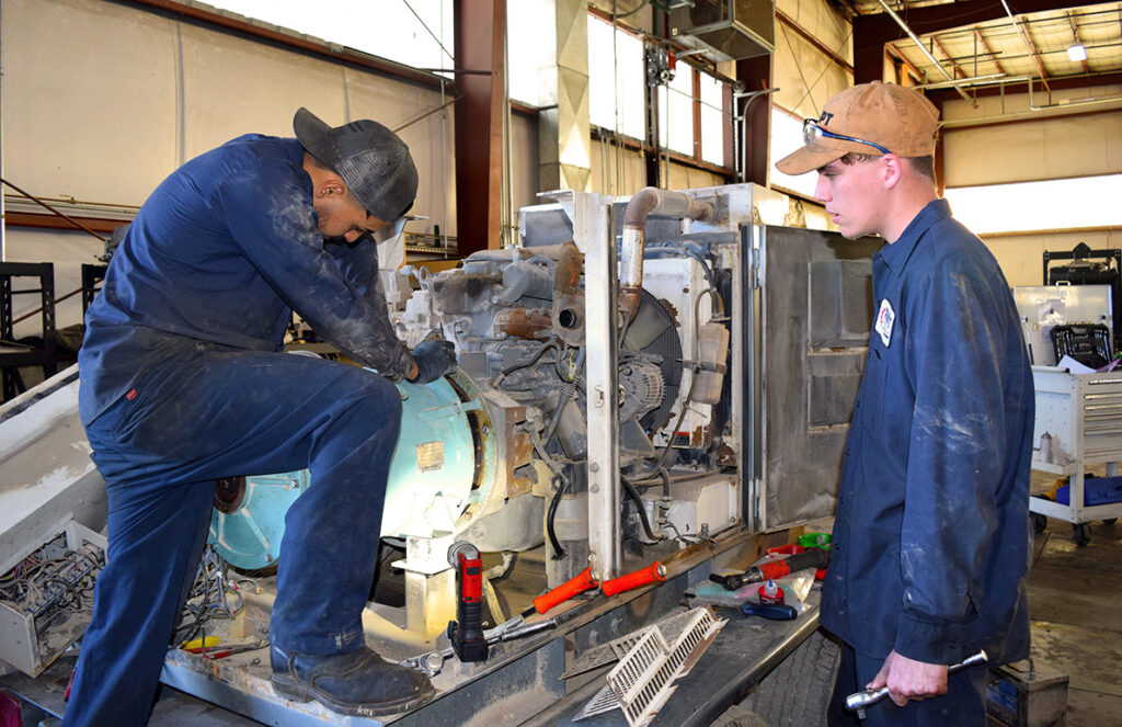 
Two mechanics are working on a large engine unit in a workshop. One mechanic is crouched over the engine, focusing intently as he uses tools to make adjustments, while the other stands nearby, observing and holding a wrench. Both are wearing dark blue work coveralls and caps, with visible dust and grease from the repair work. Various tools and parts are scattered around the engine, highlighting the hands-on, detailed nature of their task. The workshop has industrial lighting and equipment, creating a functional and rugged environment.