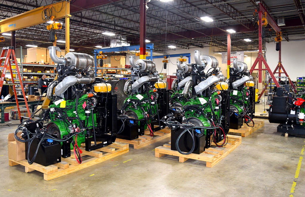 A row of large, green industrial engines sits on wooden pallets inside a spacious manufacturing facility. Each engine is equipped with prominent exhaust systems and multiple attached components, reflecting complex assembly. The facility has high ceilings with overhead cranes and an organized layout, including tool racks and workstations. The engines are prepared for further distribution or installation, showcasing the facility's capacity for high-volume production and detailed assembly work.