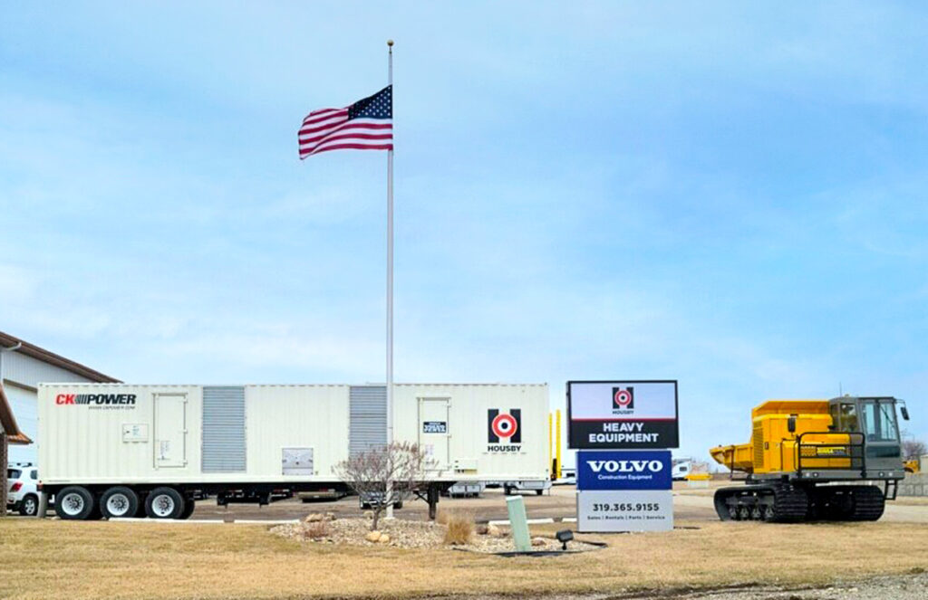 An American flag flies over a dealership displaying a large CK Power generator trailer and a yellow Terramac crawler. The dealership sign reads “Housby Heavy Equipment” and features the Volvo logo, indicating an association with construction equipment sales and service. The scene is set on a clear day with a blue sky, and the equipment is positioned prominently, showcasing the dealership's offerings for heavy-duty industrial and construction needs. The surrounding area includes a well-kept lawn and a gravel lot, adding to the professional appearance of the dealership.