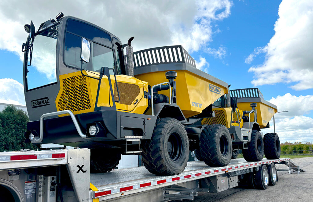 A yellow Terramac 120TW wheeled dumper, secured on a flatbed trailer, stands under a bright blue sky with scattered clouds. The dumper features a spacious cabin with large windows for visibility, and a rugged frame designed for off-road capabilities. The trailer holds two identical dumpers, highlighting their sturdy build and large, all-terrain tires suitable for various heavy-duty tasks. The Terramac branding is prominently displayed on the side.