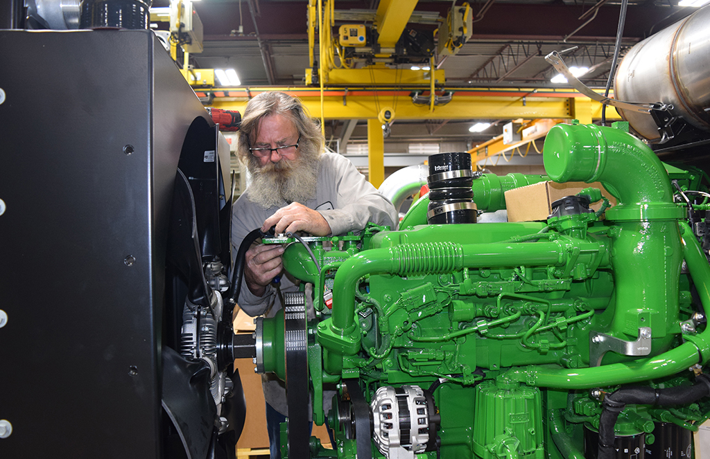 An older man with long hair and a beard is working on a large, green industrial generator in a manufacturing facility. He is adjusting a part with a tool while focused on his task. The surroundings include heavy equipment and overhead cranes in a well-lit, spacious industrial setting.