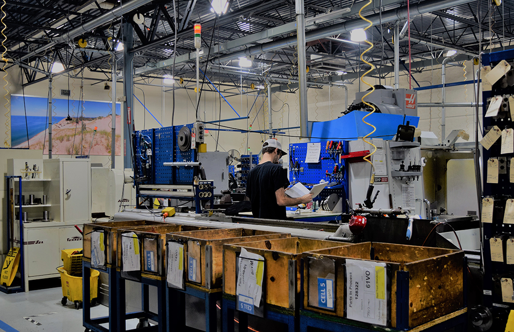 A worker in a manufacturing facility stands by a workbench, holding paperwork and inspecting machinery.