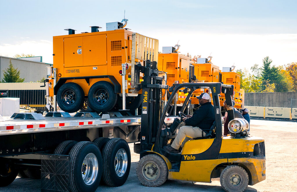 A worker operates a yellow forklift to load a portable orange generator onto a flatbed trailer. Several other similar generators are lined up in the background, indicating a busy distribution or logistics area. The setting is outdoors on a clear day, with additional equipment and storage containers visible nearby.