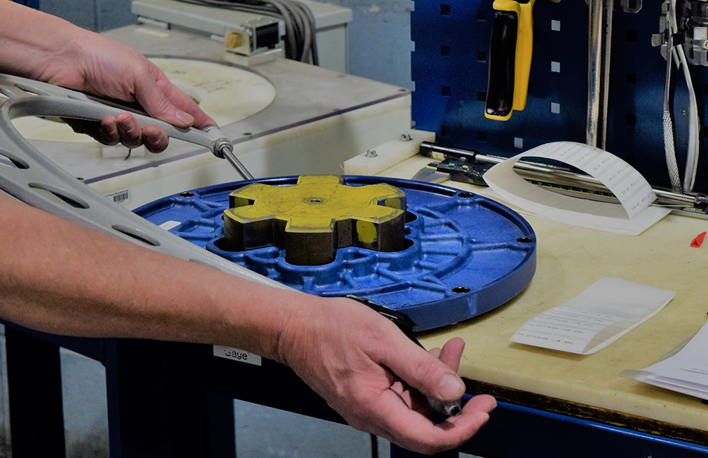 A close-up of a technician’s hands assembling or inspecting a component on a blue metal fixture in a manufacturing setting.