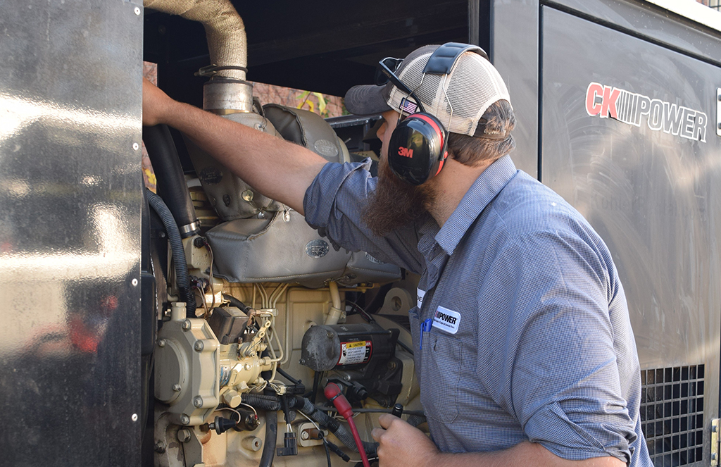 A bearded technician wearing safety earmuffs and a mesh baseball cap is performing maintenance on an off-highway power generator. He is reaching inside the generator housing, which bears the logo 'CK Power.' The technician is focused on his work, standing beside the black casing of the generator.