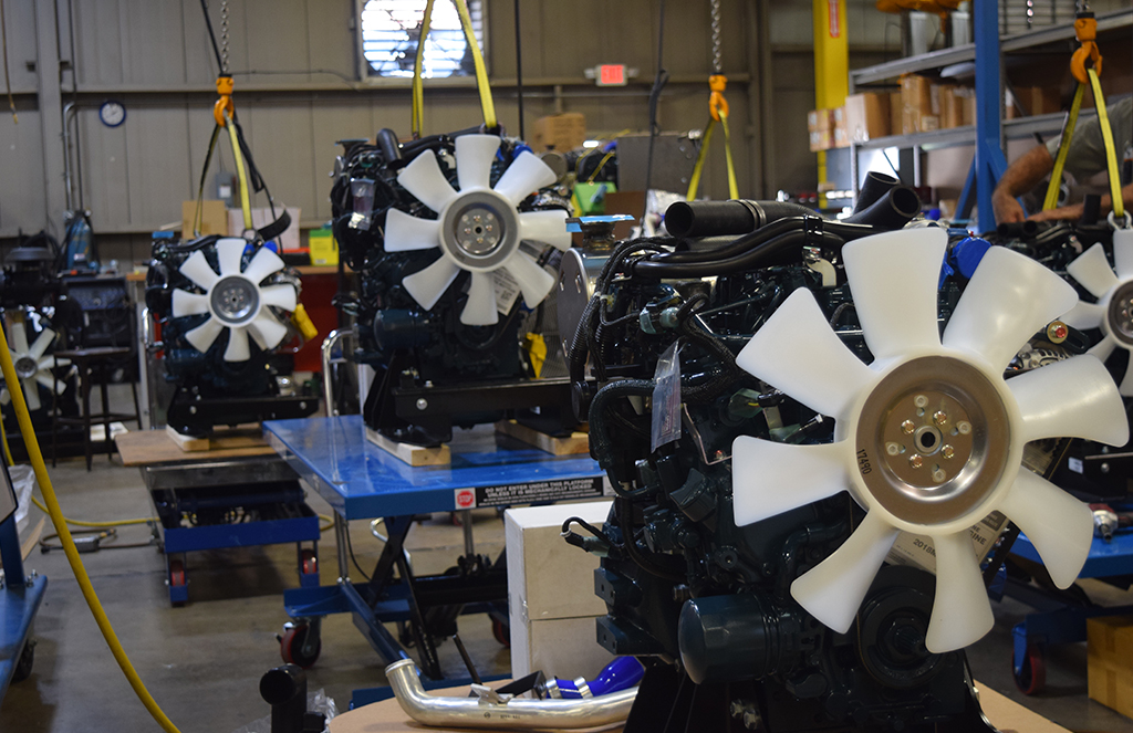 Multiple off-highway engines with large white cooling fans are positioned on blue worktables in a manufacturing facility. 